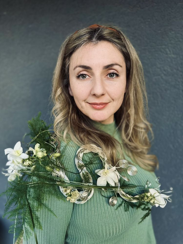 Portretul Ramonei Anghilini, în pulover verde, purtând o lucrare florală cu orhidee albe pe o structură din sârmă împletită